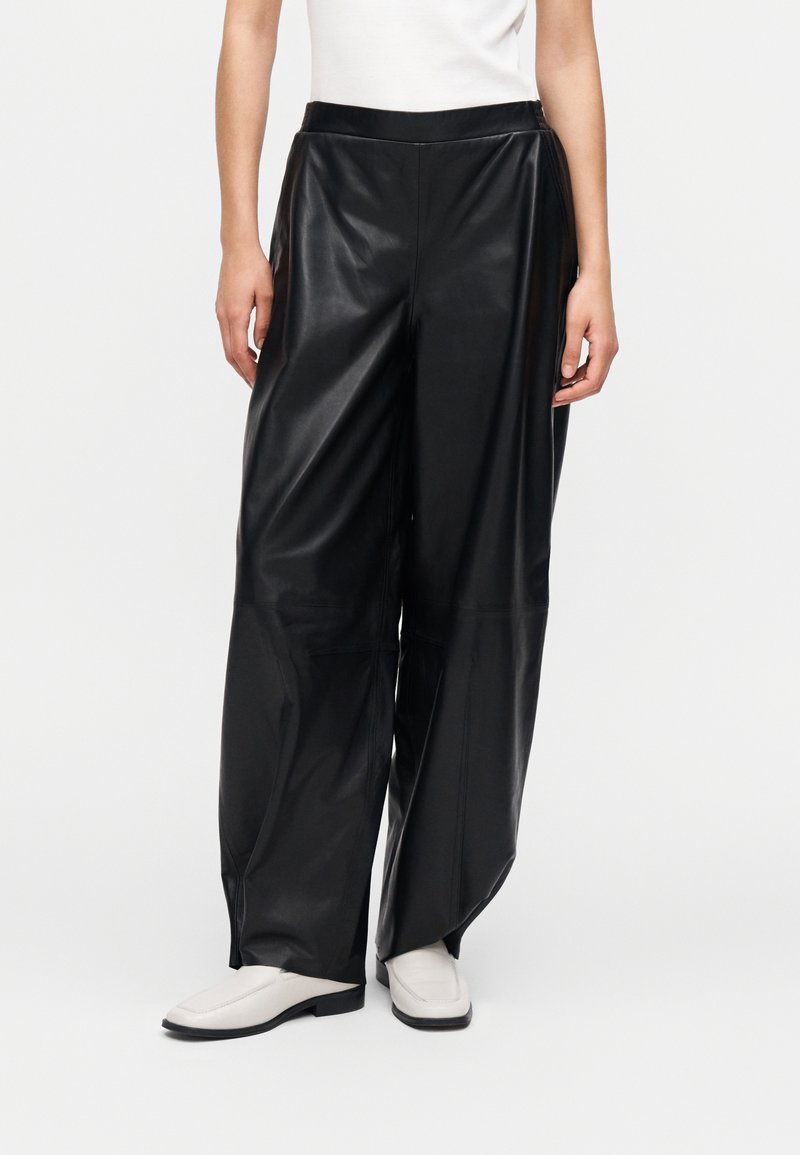 Calças de ganga largas pretas em couro falso, com cintura alta, textura suave e costuras subtis. Combinadas com sapatos deslizantes de cor clara.