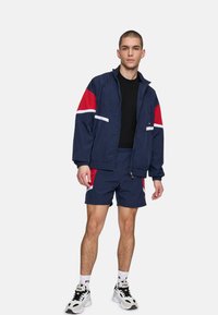 Giacca a vento navy con dettagli rossi e bianchi, abbinata a pantaloni corti coordinati. Tessuto leggero, chiusura con zip e polsini elastici. Calzature sportive casual.