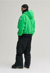 Grüne Kapuzenjacke mit elastischen Bündchen und Saum, kombiniert mit schwarzen Hosen mit Reißverschlusstaschen, getragen mit schwarzen Stiefeln.