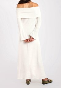 Robe longue, blanche, décolletée sur les épaules avec des manches évasées et une texture lisse, dotée d'un corsage ajusté et d'une jupe fluide. Chaussures délicates de couleur verte.