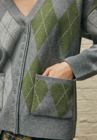 Cardigan gris avec un motif en diamant en vert foncé et gris clair, doté de boutons ronds et d'une poche avant. Texture douce et poignets côtelés.