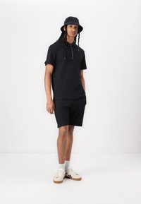 Svart kortermet polo skjorte med glidelås, matchet med svarte shorts. Brukt med en svart bøttehatt og hvite sneakers med lyse såler.