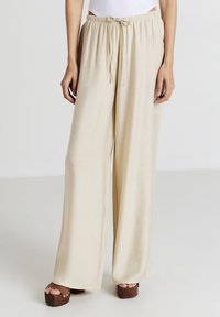 Pantalons larges beiges en tissu doux, dotés d'une taille élastique avec cordon de serrage et d'une texture lisse. Associés à des chaussures à plateforme marron.