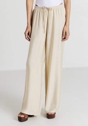 Pantaloni beige a gamba larga realizzati in tessuto morbido, con vita elasticizzata e cordino. Abbinati a scarpe con plateau marroni.