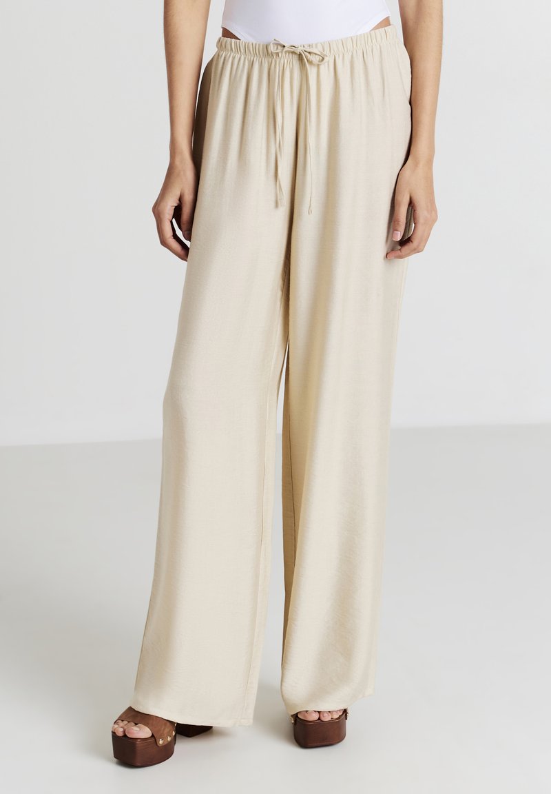 Pantalons larges beiges en tissu doux, dotés d'une taille élastique avec cordon de serrage et d'une texture lisse. Associés à des chaussures à plateforme marron.