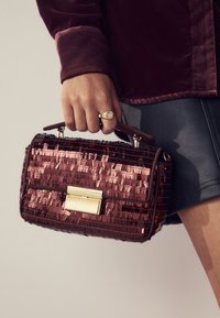 Rote Paillettenhandtasche mit strukturierter Form, goldfarbenen Details und einem oberen Griff. Das Material wirkt glänzend mit einer strukturierten Oberfläche.