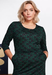 Musta mekko, jossa on geometrinen vihreä kuvio, pyöreä pääntie ja kolmivartiset hihat. Sisältää taskut ja on muotoiltu istuvaksi.
