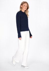 Maglione lavorato a maglia blu navy con polsini a coste, scollatura rotonda e bottoni decorativi sulla spalla, abbinato a pantaloni bianchi a gamba larga e sneaker.