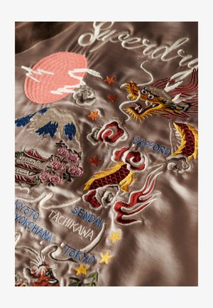 Veste bomber en satin avec des broderies colorées représentant des dragons, des fleurs et des noms de villes japonaises sur un fond brun clair.