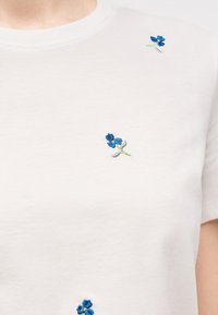 Λευκό μπλουζάκι με στρογγυλή λαιμόκοψη, διακοσμημένο με μικρά μπλε κεντημένα μοτίβα λουλουδιών διάσπαρτα στο στήθος και την περιοχή των ώμων.