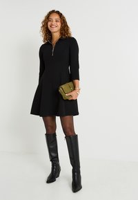 Robe noire à manches longues et col zippé, associée à des bottes noires jusqu'aux genoux. La mannequin tient un sac à main texturé vert.
