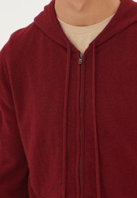 Burgundy zip-up hoodie med dragsko i huvan, mjukt tyg och ribbade ärmslut. Har en central dragkedja och en enkel design.