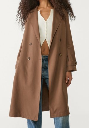 Vrouw die een lange, bruin double-breasted jas draagt over een wit cropped top met knopen en een blauwe spijkerbroek.