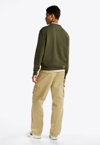 Joven de pie de espaldas con un sudadera verde olivo, pantalones cargo beiges y zapatillas blancas frente a un fondo liso.