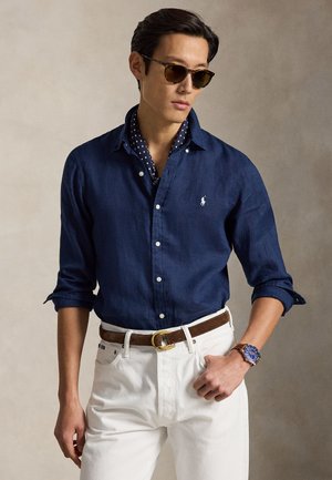 Uomo che indossa una camicia blu scuro abbottonata, pantaloni bianchi, cintura marrone, orologio blu, occhiali da sole e una sciarpa blu navy a pois intorno al collo.