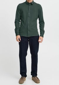 Camisa de manga larga abotonada de color verde oscuro, tela ligera, cuello clásico, combinada con pantalones azul oscuro y zapatos de ante marrón.