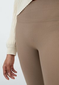 Leggings marrones de canalé con cintura alta, fabricados en un material elástico. Un suéter texturizado color crema parcialmente visible por encima.