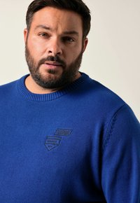 Blauer gestrickter Pullover mit Rundhalsausschnitt, der über eine strukturierte Naht und drei dunkelblaue gestickte Patches auf der Brust verfügt.