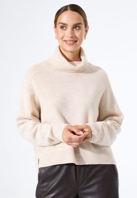 Beige gestrickter Pullover mit einem Rollkragen, gerippten Bündchen und Saum. Weiche Textur und lockere Passform, kombiniert mit dunklen Lederhosen.