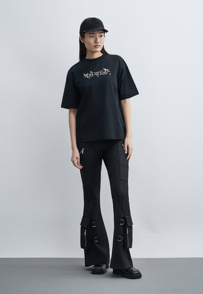 Νεαρή γυναίκα που φοράει μαύρο καπέλο μπέιζμπολ, μαύρο μπλουζάκι Off-White, μαύρο παντελόνι cargo με αγκράφες και μαύρα παπούτσια, στέκεται μπροστά σε γκρι φόντο.