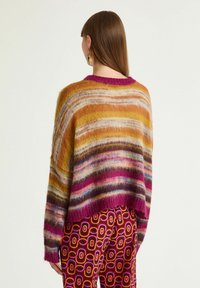 Donna con capelli castani lisci, che indossa orecchini d'oro, un maglione a righe morbido e multicolore e pantaloni con motivi geometrici, vista da dietro.