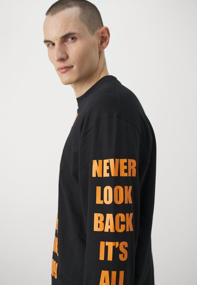 T-shirt noir à manches longues avec un texte orange vif sur la manche et à l'avant. Le matériau semble doux ; le design est simple et moderne.