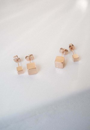 Zwei Paare roségoldene quadratische Ohrstecker, ein Paar mit größeren und das andere mit kleineren Würfeln, auf einer weißen Oberfläche dargestellt.