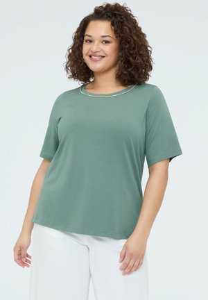 Smilende kvinde med krøllet hår iført en grøn kortærmet top med perledekoreret halsudskæring og hvide bukser, stående mod en ensfarvet baggrund.