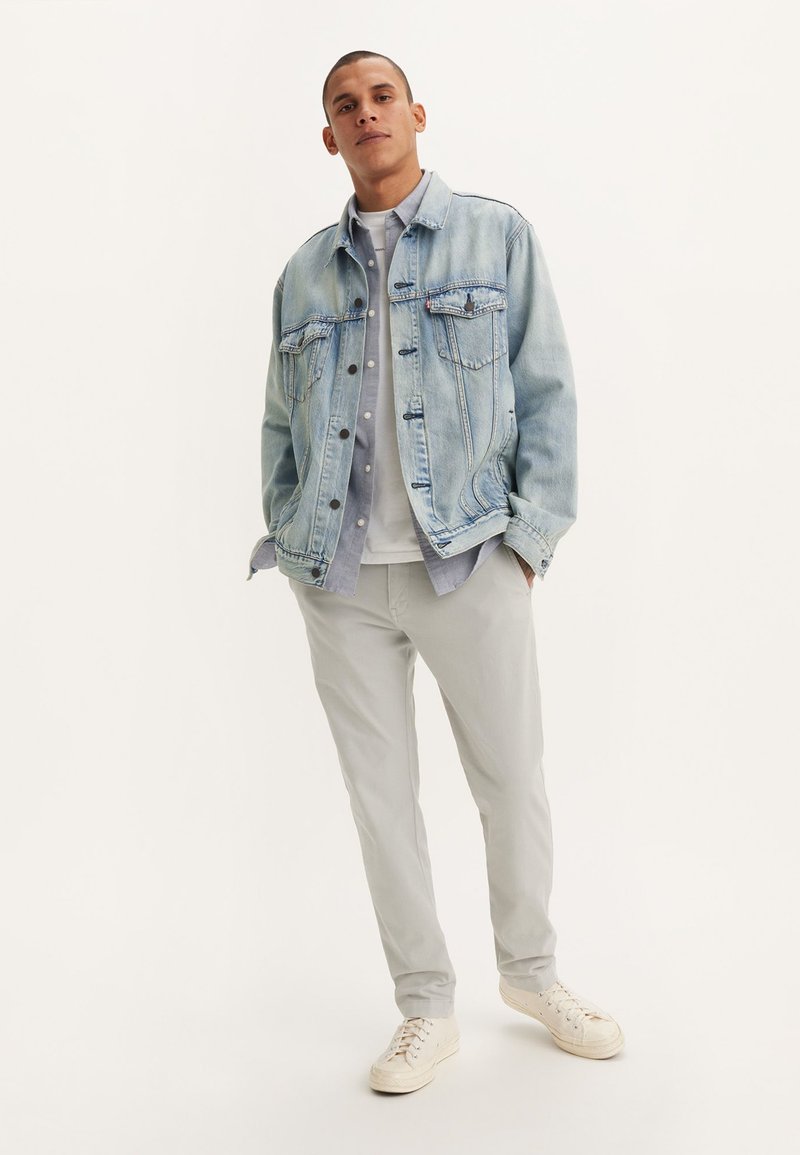 Chaqueta de mezclilla azul clara con dos bolsillos en el pecho, camisa de botones gris debajo, pantalones grises y zapatillas blancas. Diseño simple, corte relajado.
