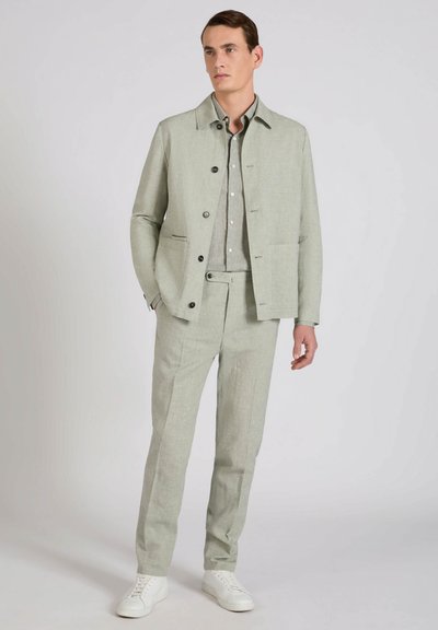 Hombre con chaqueta abotonada gris claro, pantalones a juego, camisa gris y zapatillas blancas, de pie con una mano en el bolsillo.