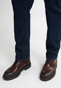 Botas de cuero marrón con punta redondeada, suela de goma negra y cordones. Combinadas con unos vaqueros de mezclilla azul oscuro, mostrando un estilo casual.