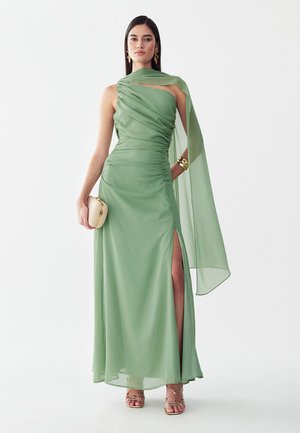 Vrouw in een lichtgroene, asymmetrische jurk met gedrapeerde stof, zijsplit, gouden sieraden, sandalen met bandjes en met een kleine beige clutch in de hand.