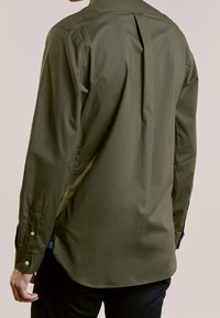 Olivgrön skjorta med långa ärmar, bakfald, knappmanchetter och rundad nederkant. Tillverkad av ett slätt tyg med en minimalistisk design.