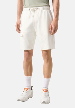 Mężczyzna ubrany w białe bermudy cargo do kolan z wiązanym paskiem w talii, do tego białe skarpetki i białe trampki z pomarańczowymi sznurowadłami.