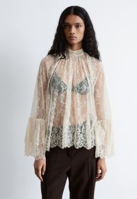 Spitzenoberteil in Creme mit ausgestellten Ärmeln und gewelltem Saum, mit einem hohen Kragen und transparentem Overlay. Braune Hosen vervollständigen das Outfit.