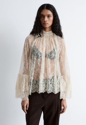 Spitzenoberteil in Creme mit ausgestellten Ärmeln und gewelltem Saum, mit einem hohen Kragen und transparentem Overlay. Braune Hosen vervollständigen das Outfit.