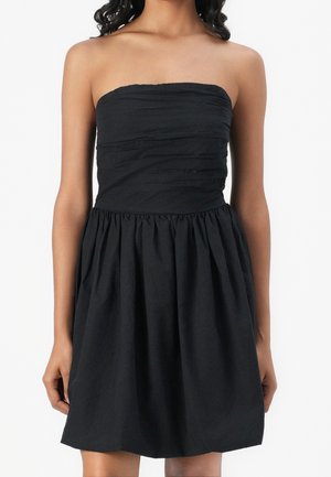 Czarna sukienka bez ramiączek z marszczonym biustem i marszczonym dołem, noszona przez osobę o średnio-ciemnym odcieniu skóry na białym tle.