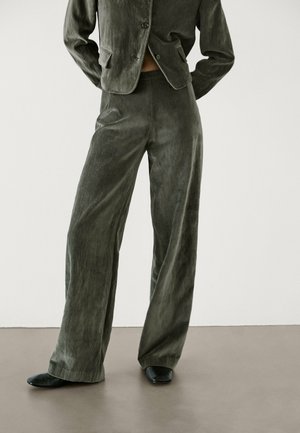 Modèle portant un pantalon en velours large couleur olive, une veste courte assortie et des chaussures noires à bouts pointus, debout sur un sol gris.