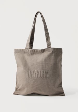 Beige lerretveske med to håndtak og "DRYKORN" preget på forsiden, står oppreist på en enkel hvit bakgrunn.