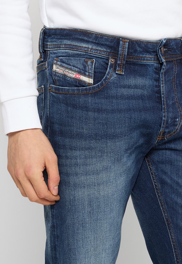 Jean en denim bleu foncé avec une texture délavée, présentant une petite étiquette de poche avant avec le label "Diesel" et des coutures orange contrastées.