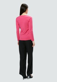 Langärmliges, figurbetontes pinkes Oberteil mit Rundhalsausschnitt, kombiniert mit schwarzen, weiten Hosen und High Heels, von hinten betrachtet.