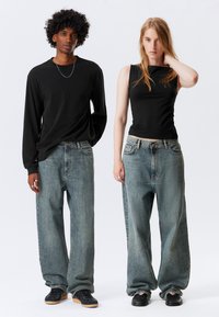 Δύο μοντέλα φορούν υπερμεγέθη τζιν: το ένα με μακρυμάνικο μαύρο πουκάμισο, το άλλο με εφαρμοστό μαύρο τοπ χωρίς μανίκια. Τα παπούτσια παρουσιάζουν αντιθετικά σχέδια.