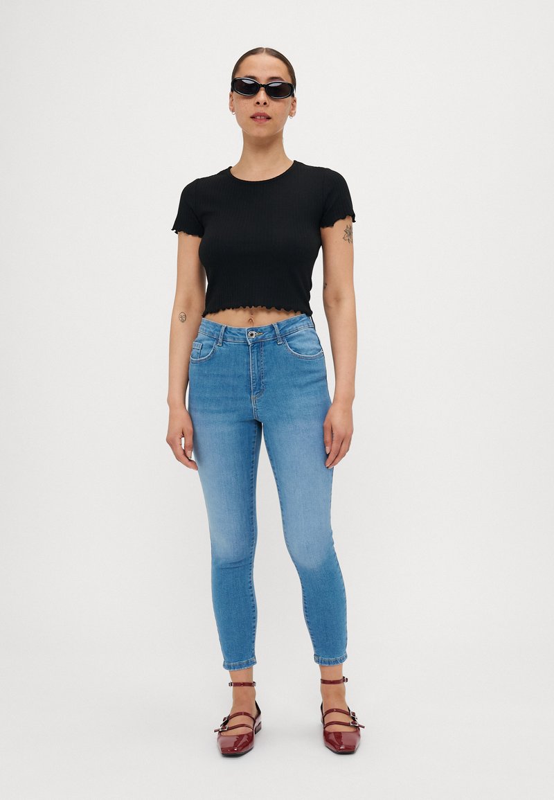 Schwarzes, geripptes Cropped-Top mit gerafften Kanten, hellblaue, figurbetonte Jeans und rot lackierte Lederschuhe mit Riemen und Schnallen. Schwarze Sonnenbrille getragen.