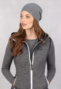 Grijze gebreide muts met geribbelde structuur. Draagt een grijze bodywarmer met rits, voorzien van een logo en witte accenten. Haar valt los over de schouders.