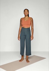 Kvinne med fletter som har på seg en oransje crop top og grå vide bukser, står barbeint på en beige yogamatte mot en ensfarget bakgrunn.