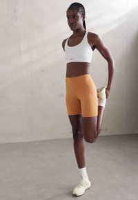 Conjunto atlético que incluye un sujetador deportivo blanco y pantalones cortos de tiro alto naranja. Combinado con calcetines blancos y zapatillas crema. Pose de estiramiento.