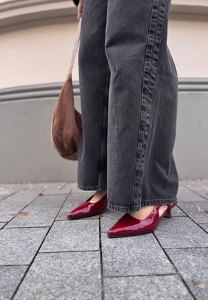 Rode gepatineerde leren slingback hakken met een spitse neus en een kleine hak, gecombineerd met brede grijze denim broeken, geplaatst op een stenen stoep.