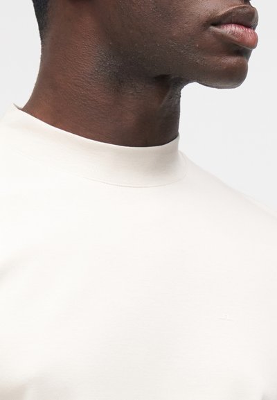 Prim-plan al unei persoane îmbrăcate cu un tricou alb simplu cu gât rotund, arătând gâtul, linia maxilarului și partea superioară a pieptului.