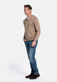Beige gebreide trui met V-hals, gedragen over een wit shirt, gecombineerd met lichtblauwe denim jeans en donkerbruine suède enkellaarsjes.