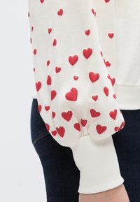 Λευκό φούτερ με ριμπ τελείωμα, διακοσμημένο με ανάγλυφες κόκκινες καρδιές σε δ ιάφορα μεγέθη διάσπαρτες στο ύφασμα, ελαστικές μανσέτες.